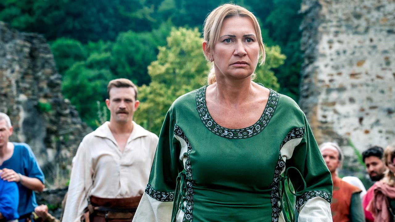 Biggi Obergschwendter  in grünem Mittelaltergewand schreitet vor einer Burgkulisse durch eine versammelte Gruppe.