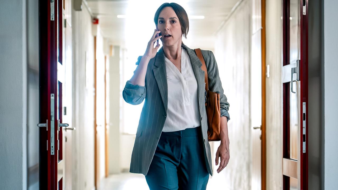 Patrizia Specht in einer weißen Bluse mit Blazer telefoniert eilig in einem langen, hellen Flur.