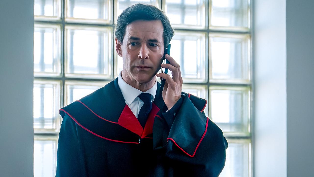 Staatsanwalt Dr. Christian Windisch im Talar telefoniert am Handy angespannt vor einer großen Fensterfront.
