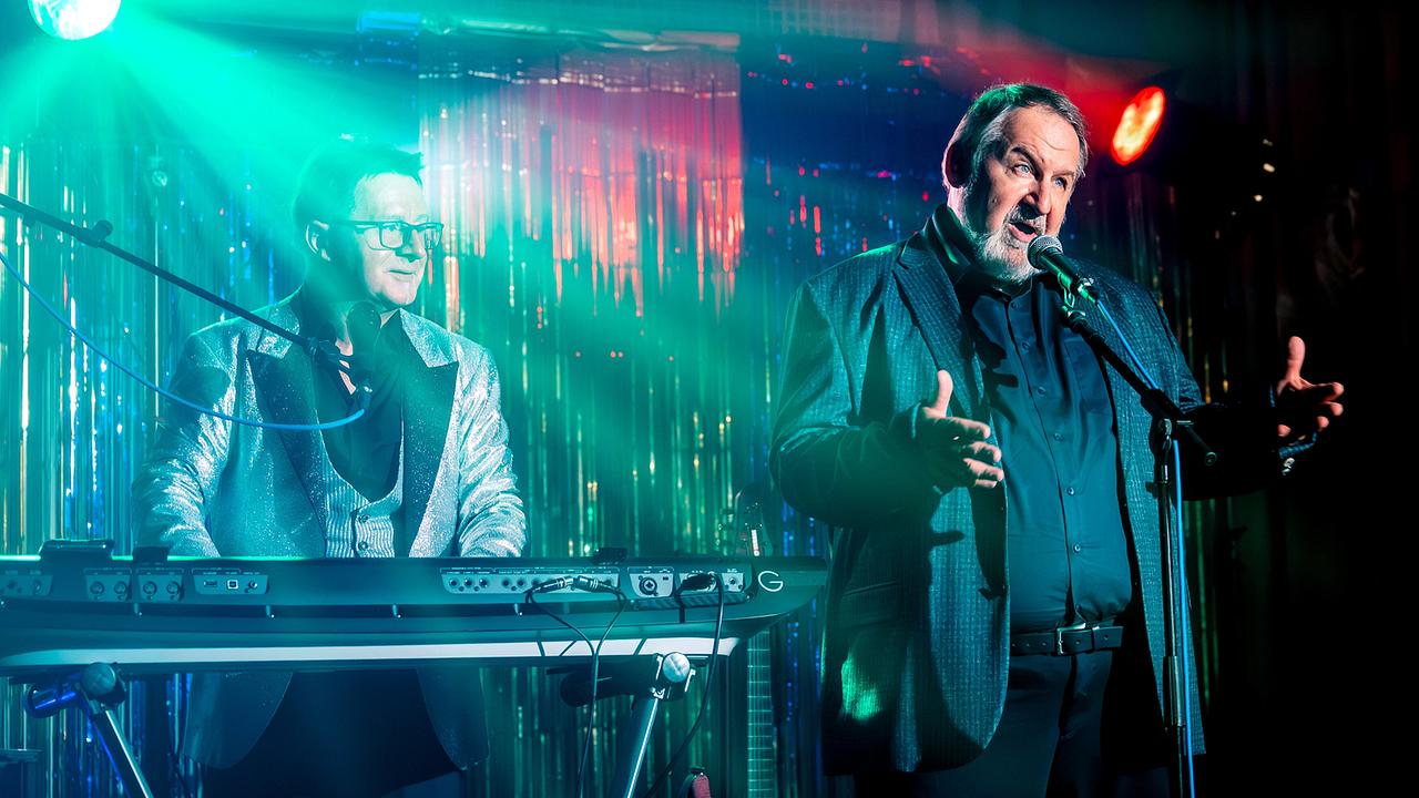 © ORF/Gebhardt Productions/Stefanie Leo Otto Komosny am Mikrofon tritt mit einem Keyboarder unter farbigem Bühnenlicht in einem Nachtlokal auf.