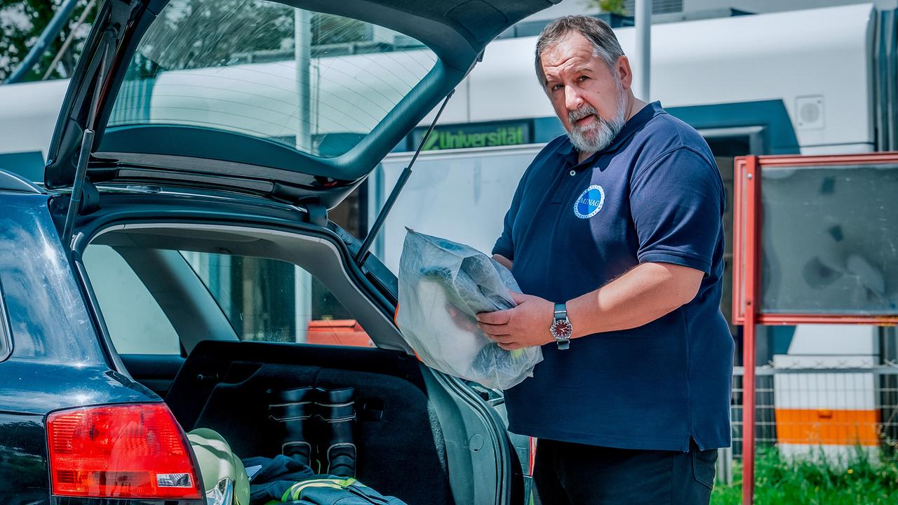 © ORF/Gebhardt Productions/Stefanie Leo Otto Komosny in MINAG-Arbeitskleidung lädt Ausrüstung aus dem Kofferraum eines Autos vor einem Gebäude aus.