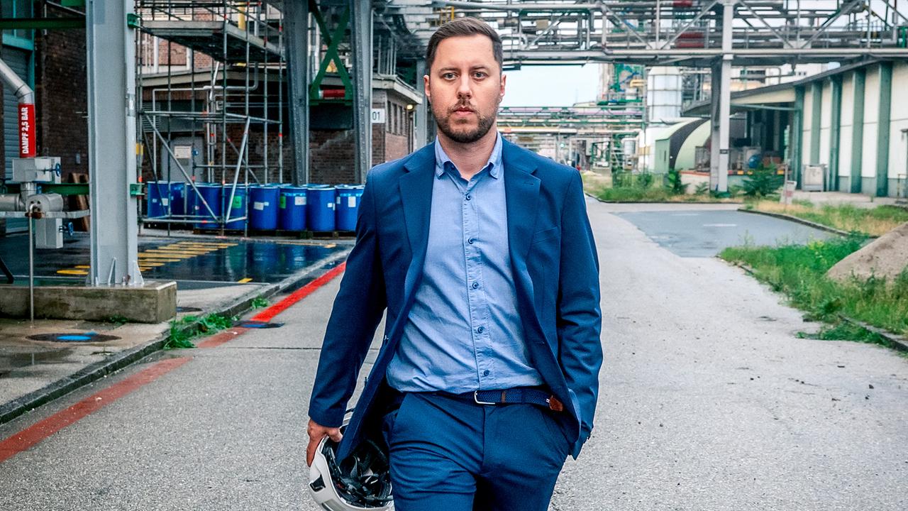 © ORF/Gebhardt Productions/Stefanie Leo Pirmin Zissner im Anzug geht mit einem Schutzhelm in der Hand durch das Gelände einer Industrieanlage.