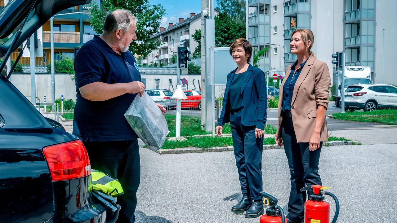 © ORF/Gebhardt Productions/Stefanie Leo Joe Haizinger und Biggi Obergschwendter sprechen mit Otto Komosny in MINAG-Arbeitskleidung neben einem geöffneten Auto, während Feuerlöscher daneben auf der Parkfläche stehen.