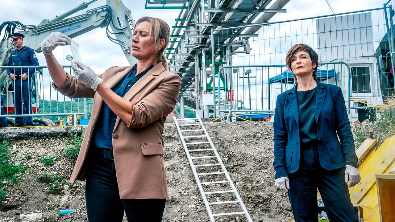 © ORF/Gebhardt Productions/Stefanie Leo Biggi Obergschwendter sichert auf einer Baustelle ein kleines Beweisstück, während Joe Haizinger daneben steht und ein Polizist im Hintergrund zu sehen ist.