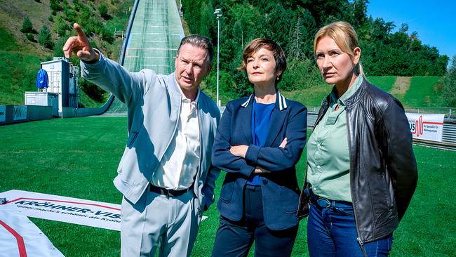 © ORF/Gebhardt Productions/Stefanie Leo Vor einer Skisprungschanze im Grünen zeigt Mark Kröhner, der neben Joe Haizinger und Biggi Obergschwendter steht, in eine Richtung.