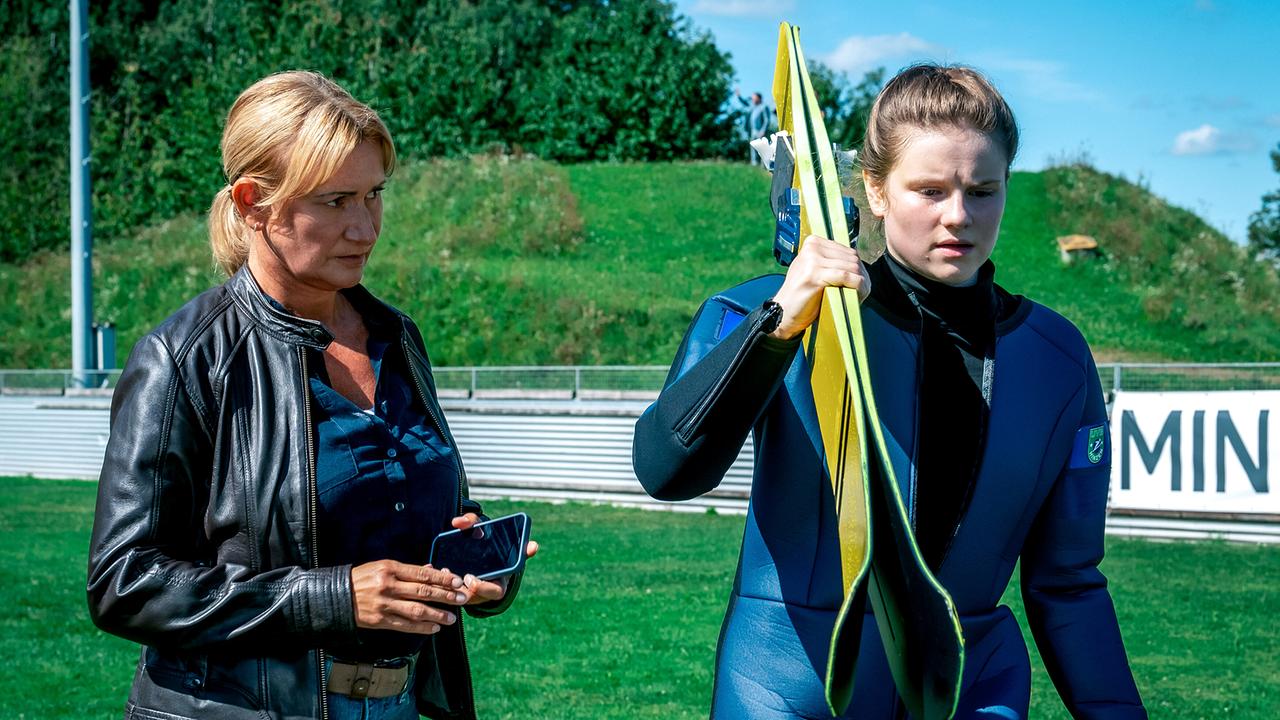 Biggi Obergschwendter in Lederjacke begleitet im Sommer eine junge Skispringerin, die ihre Skier trägt.