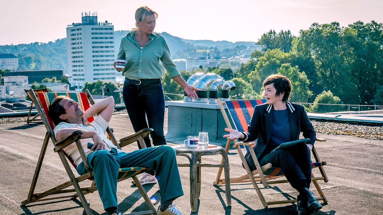 Dr. Richie Vitek, Biggi Obergschwendter und Joe Haizinger sitzen und stehen entspannt auf einer Dachterrasse in Liegestühlen und besprechen sich bei Kaffee.