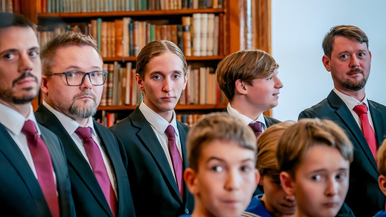 Mehrere festlich gekleideten Männer und Buben, unter ihnen Enno Tomberg, schauen in einem Raum mit großer Bücherwand aufmerksam in dieselbe Richtung.