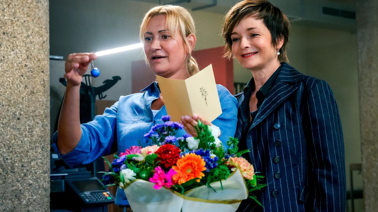 Biggi steht mit einem Blumenstrauß und Karte vor einer Tür und hält einen Schlüsselanhänger hoch, während Joe lächelnd daneben steht.