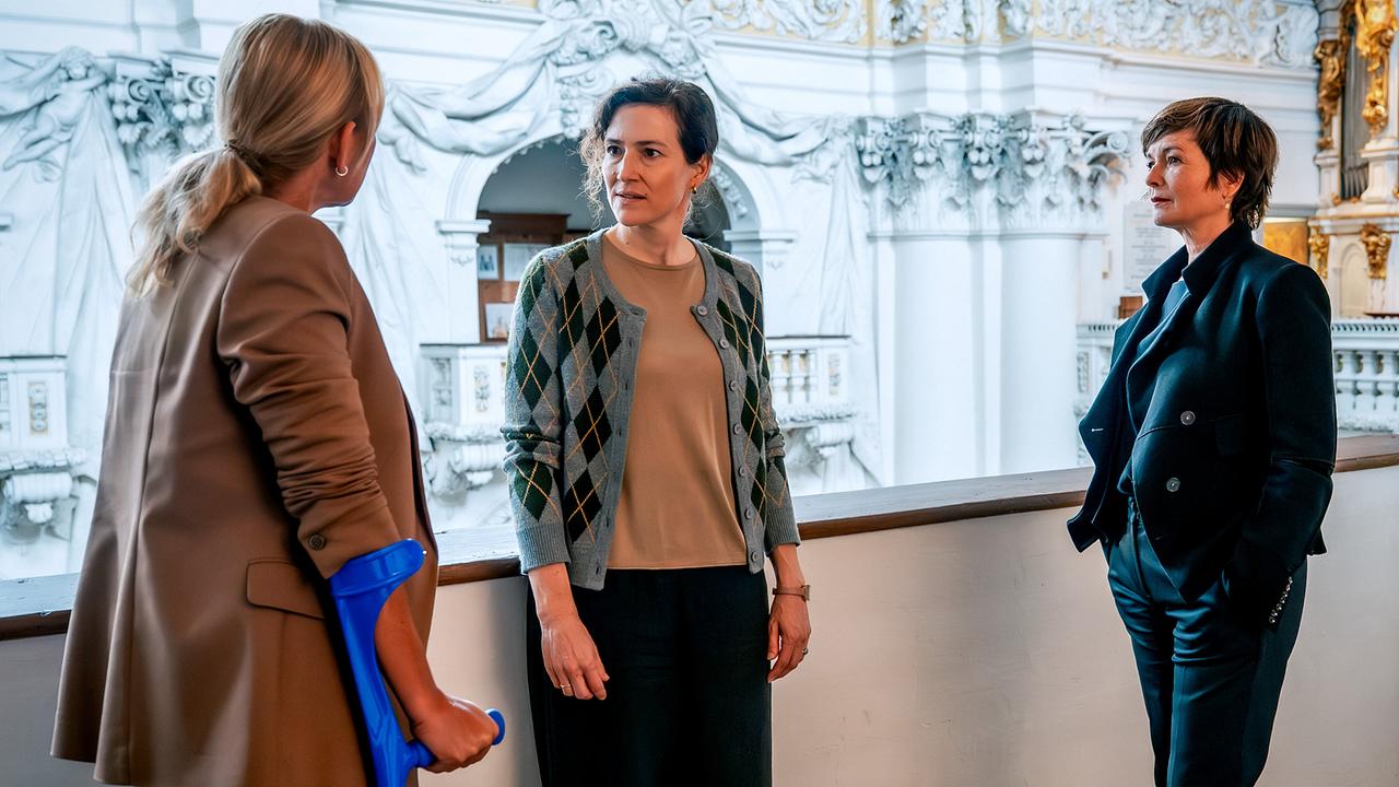 Biggi mit Krücken und Joe befragen Isolde Überbacher in einem prachtvoll ausgestatteten Gebäude.