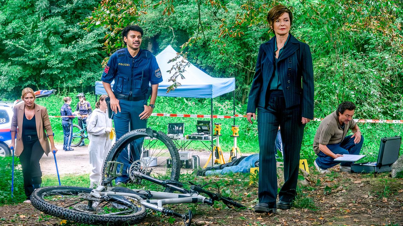 An einem abgesperrten Waldstück untersuchen Biggi, Joe und Dr. Richie Vitek sowie weitere Polizisten und die Spurensicherung einen Tatort mit Fahrrad und Leiche.