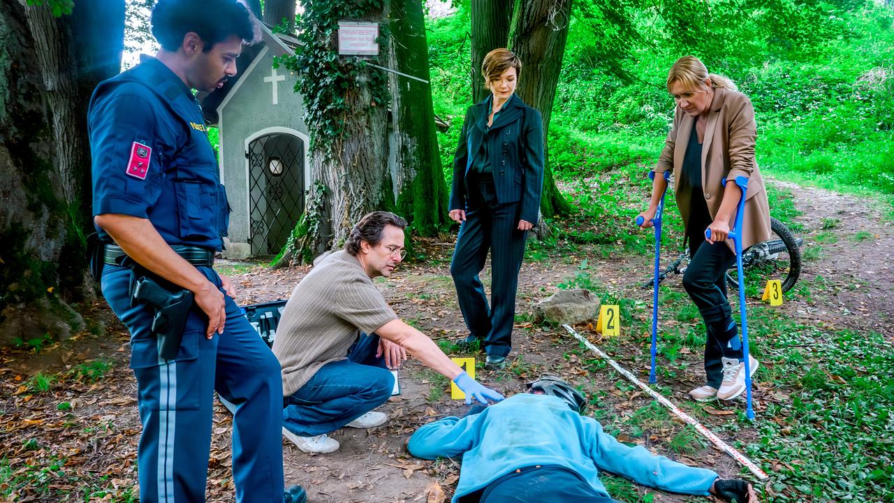 Polizist Kevin Schneeberger, Dr. Richie Vitek, Joe und Biggi untersuchen die Leiche des Radfahrers in einem Waldstück.