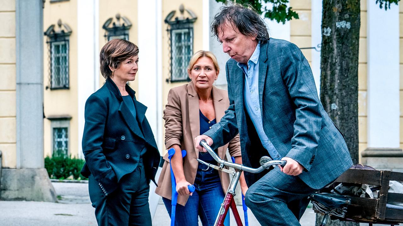 Joachim "Habi" Haberstetter fährt mit seinem alten Fahrrad an Joe und Biggi mit Krücken vor einem historischen Gebäude vorbei.