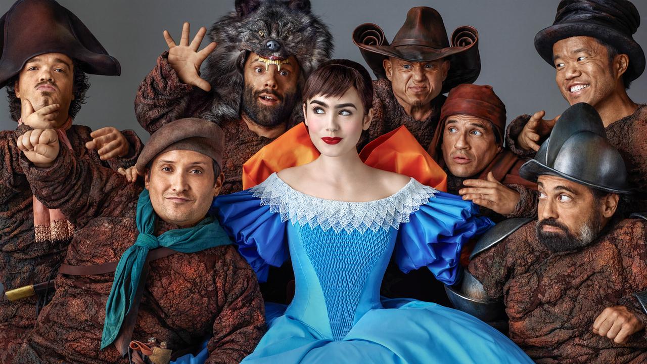 Lily Collins als Schneewittchen in einem bezaubernden hellblauen Kleid, umringt von sieben, braun gekleideten, freundlichen Männern umgeben, lächelt sie verschmitzt 