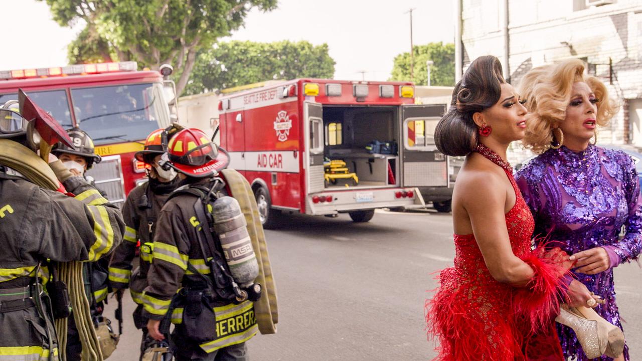 Zwei glamurös gekleidete Drag Queens stehen auf der Straße und blicken aufgeregt in dieselbe Richtung, hinter ihnen Feuerwehrmänner und zwei Einsatzfahrzeuge