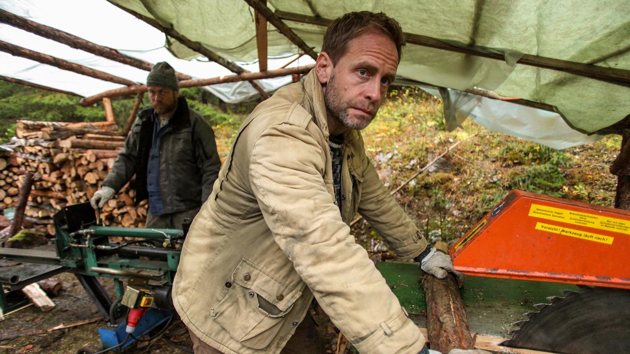 Veit Schindler arbeiten im Freien an einer Sägemaschine unter einem provisorischen Dach, hinter ihm ist ein weiterer Arbeiter zu sehen.