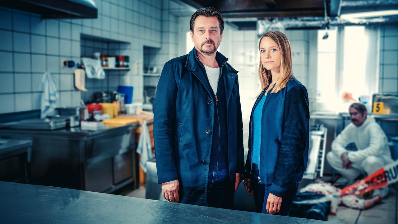 Sascha Bergmann und Anni Sulmtaler stehenan einem Tatort in einer Großküche, während im Hintergrund ein Forensiker arbeitet.