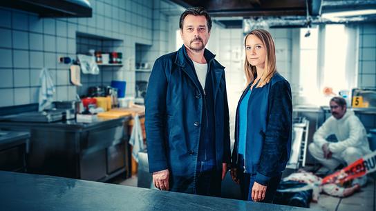 Sascha Bergmann und Anni Sulmtaler stehenan einem Tatort in einer Großküche, während im Hintergrund ein Forensiker arbeitet.