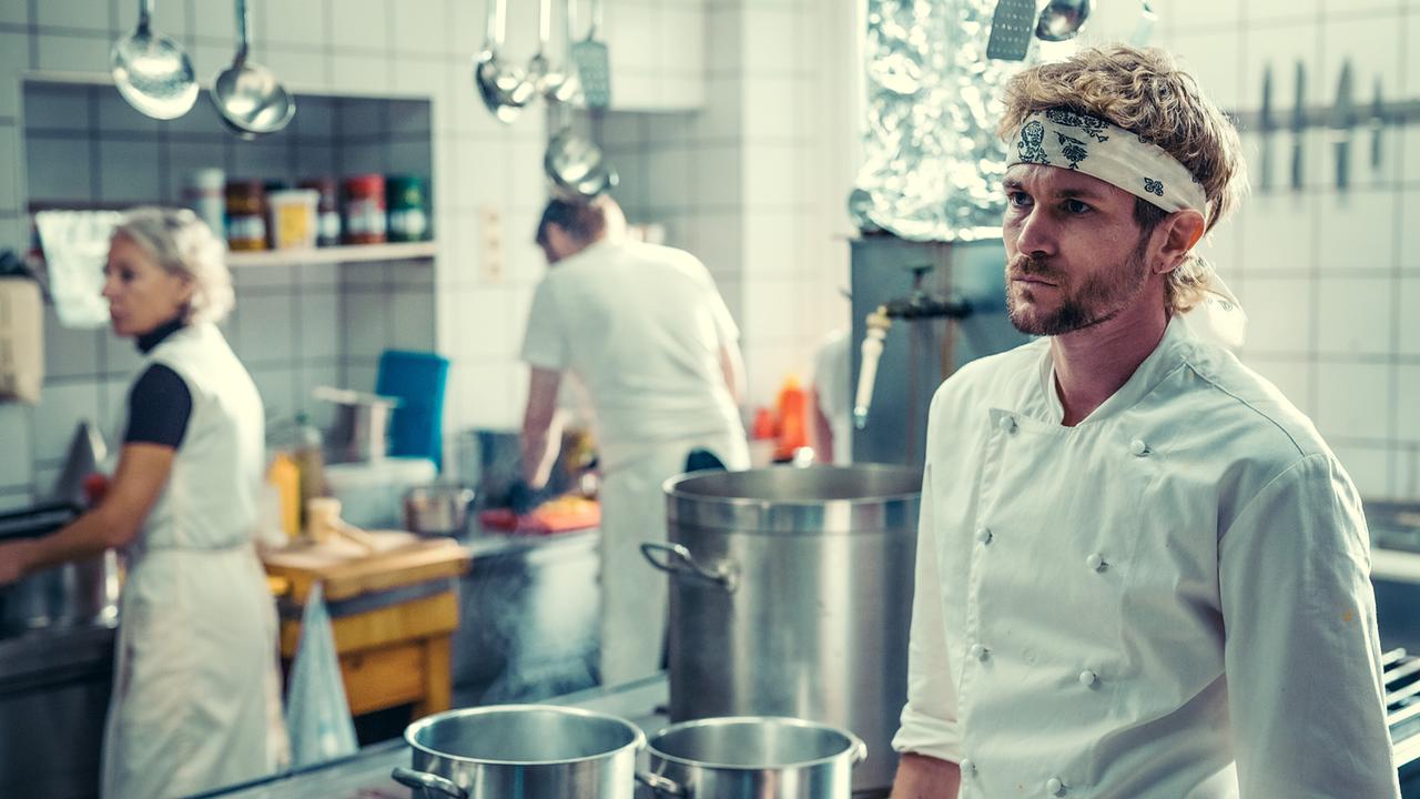 Koch Mario Höfler steht in einer geschäftigen Restaurantküche zwischen großen Töpfen und Küchenutensilien.