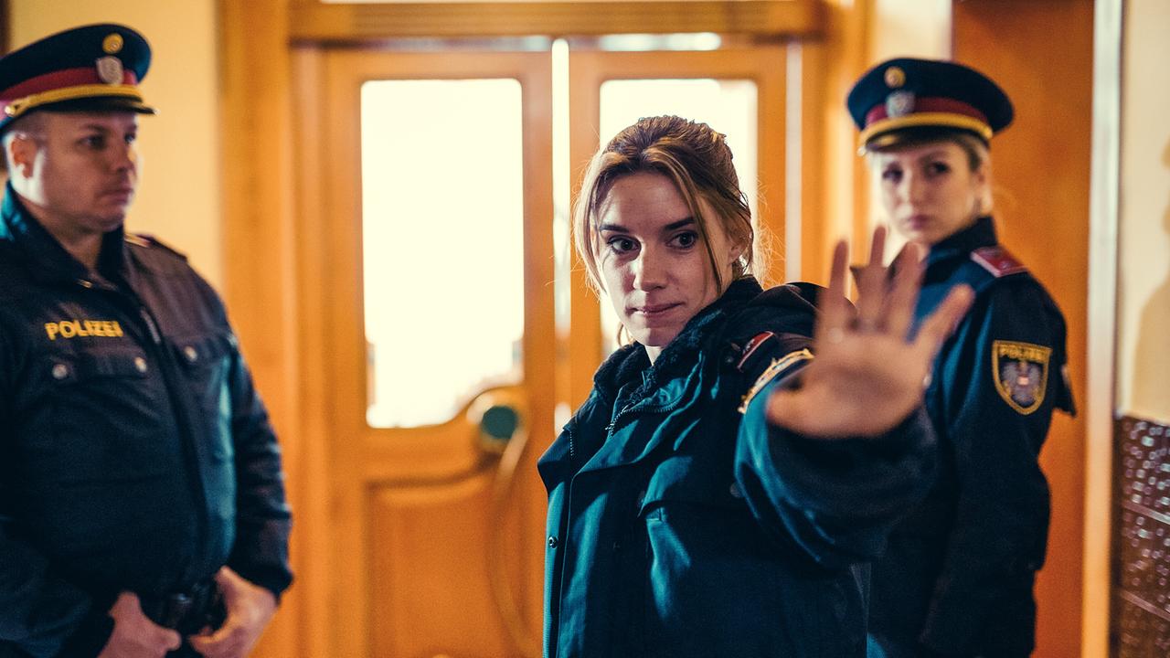 Polizistin Lena Herzog im Eingangsbereich eines Gebäudes hebt die Hand, um jemanden am Betreten eines Gebäudes zu hindern, nenem ihr zwei weitere Polizisten.