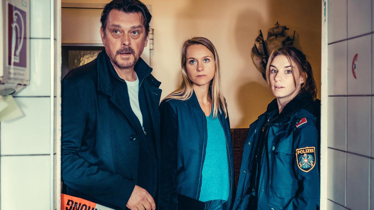 Sascha Bergmann, Anni Sulmtaler und Lena Herzog stehen hinter einer Polizeiabsperrung in einem Flur.