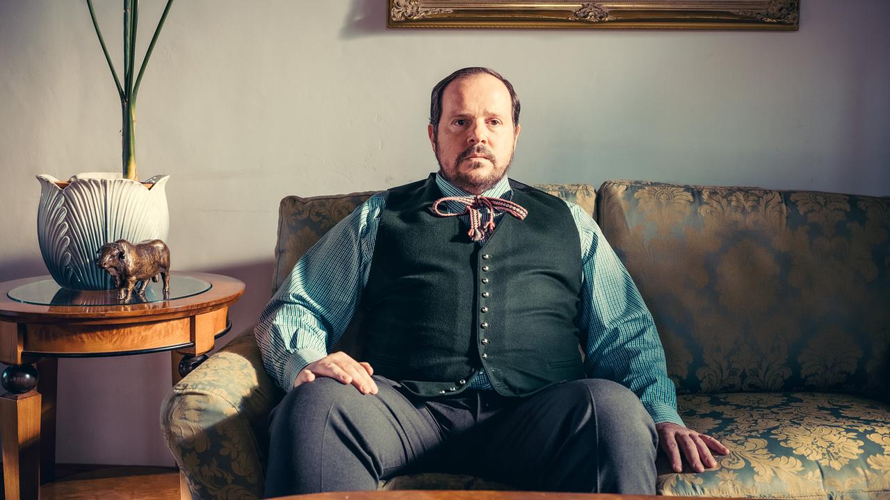 Karl Höfler  sitzt in traditioneller Kleidung auf einem Sofa in einem Wohnzimmer.