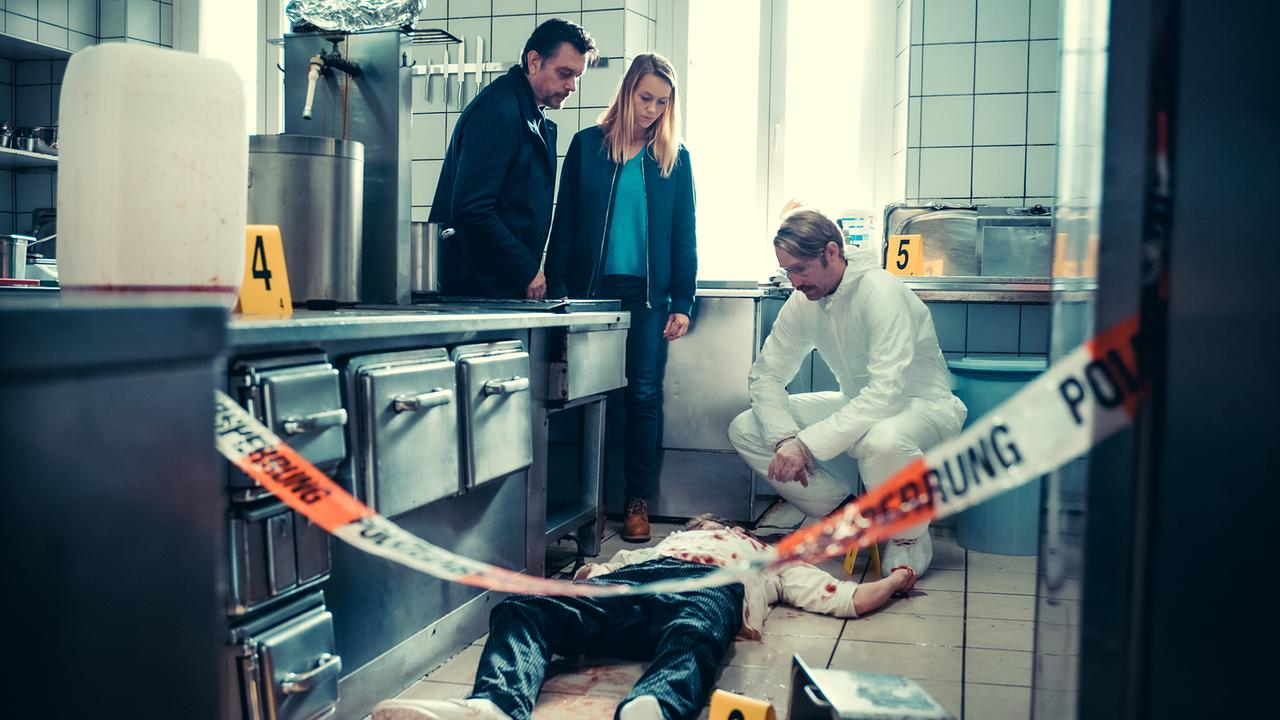 Sascha Bergmann, Anni Sulmtaler und Bernd Kofler untersuchen eine Blutspur und eine am Boden liegende Person in einer abgesperrten Küche.
