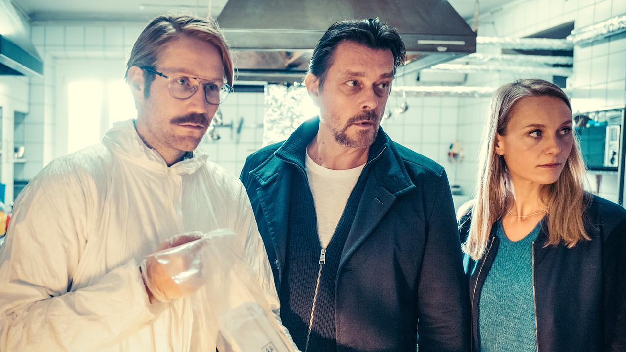 Bernd Kofler, Sascha Bergmann und Anni Sulmtaler stehen in einer Großküche, während Kofler davon einen Beweisbeutel hält.