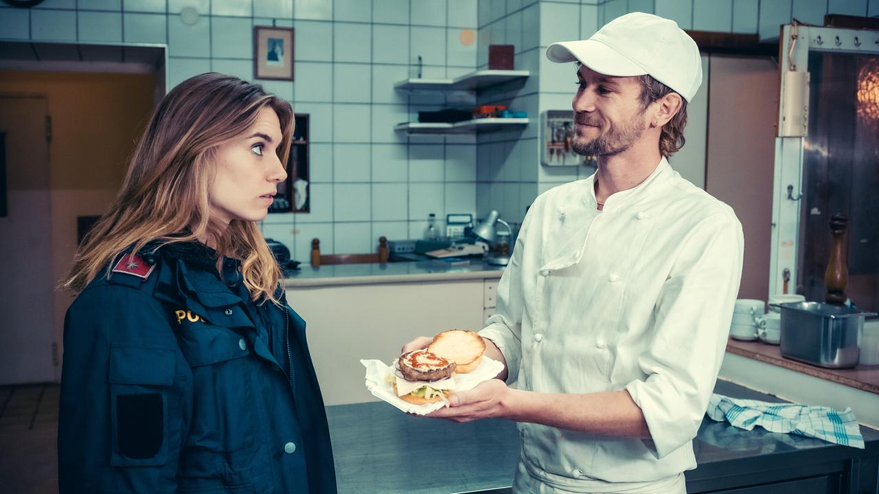 Koch Mario Höfler reicht Polizistin Lena Herzog einen Burger verziert mit einem Ketchup-Herz in einer Großküche.