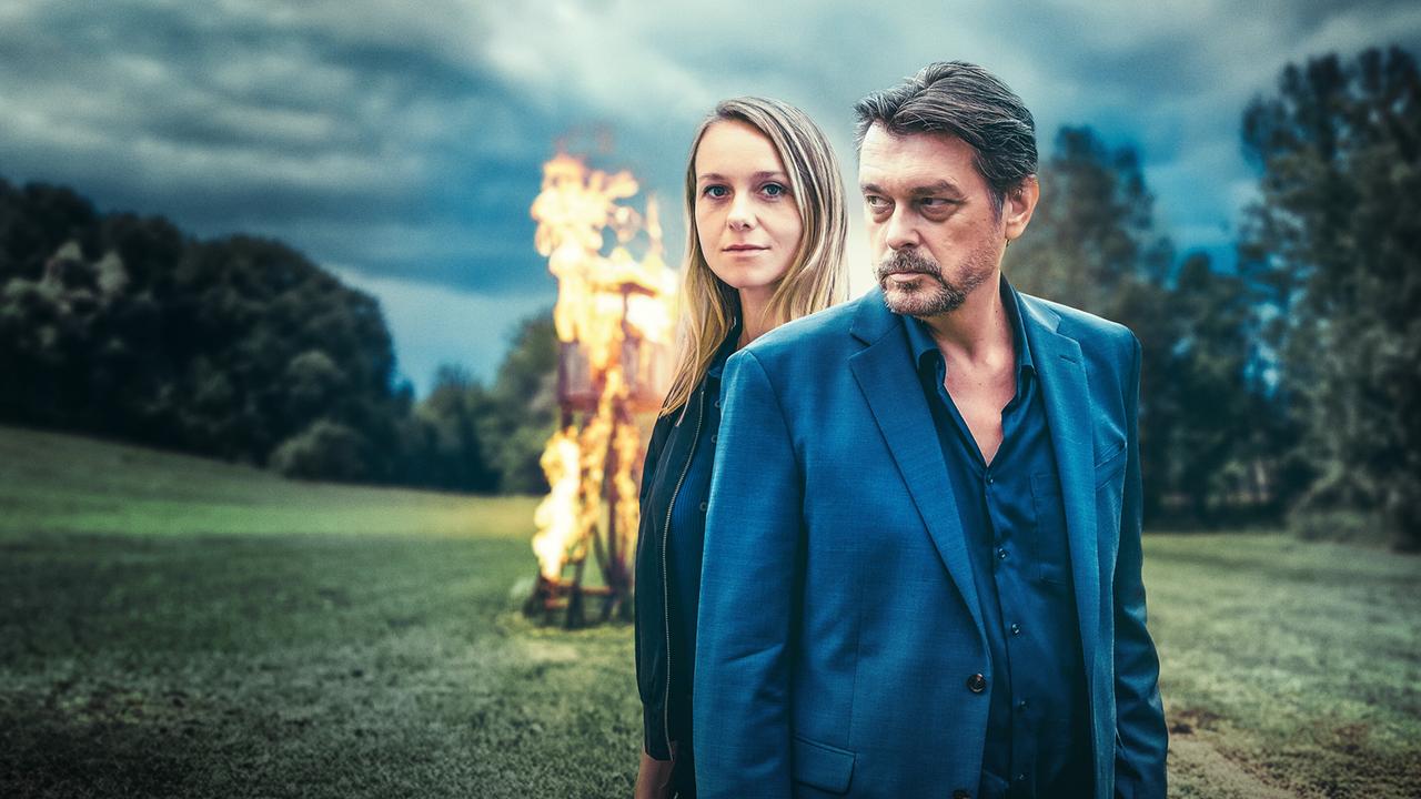 Anna Unterberger und Hary Prinz stehen auf einer Wiese, im Hintergrund brennt eine Holzstruktur.