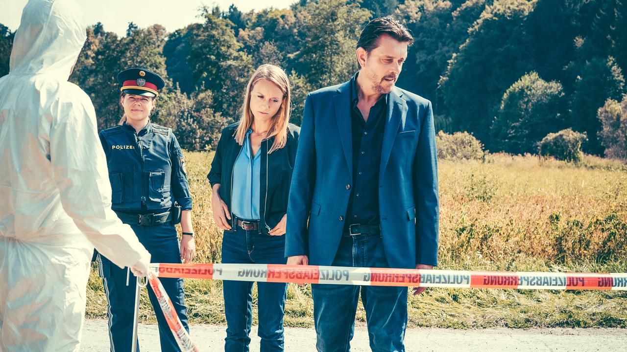 Eine Polizistin, Anna Unterberger und Hary Prinz Polizisten stehen am Tatort an einer abgesperrten Wiese, vor ihnen eine Person im Schutzanzug.