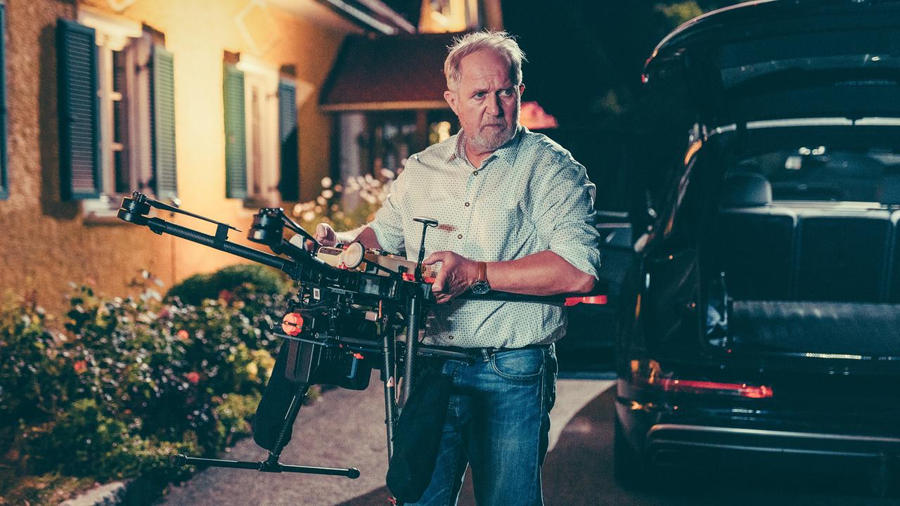 Harald Krassnitzer hält eine große Drohne, neben einem Auto bei Nacht.