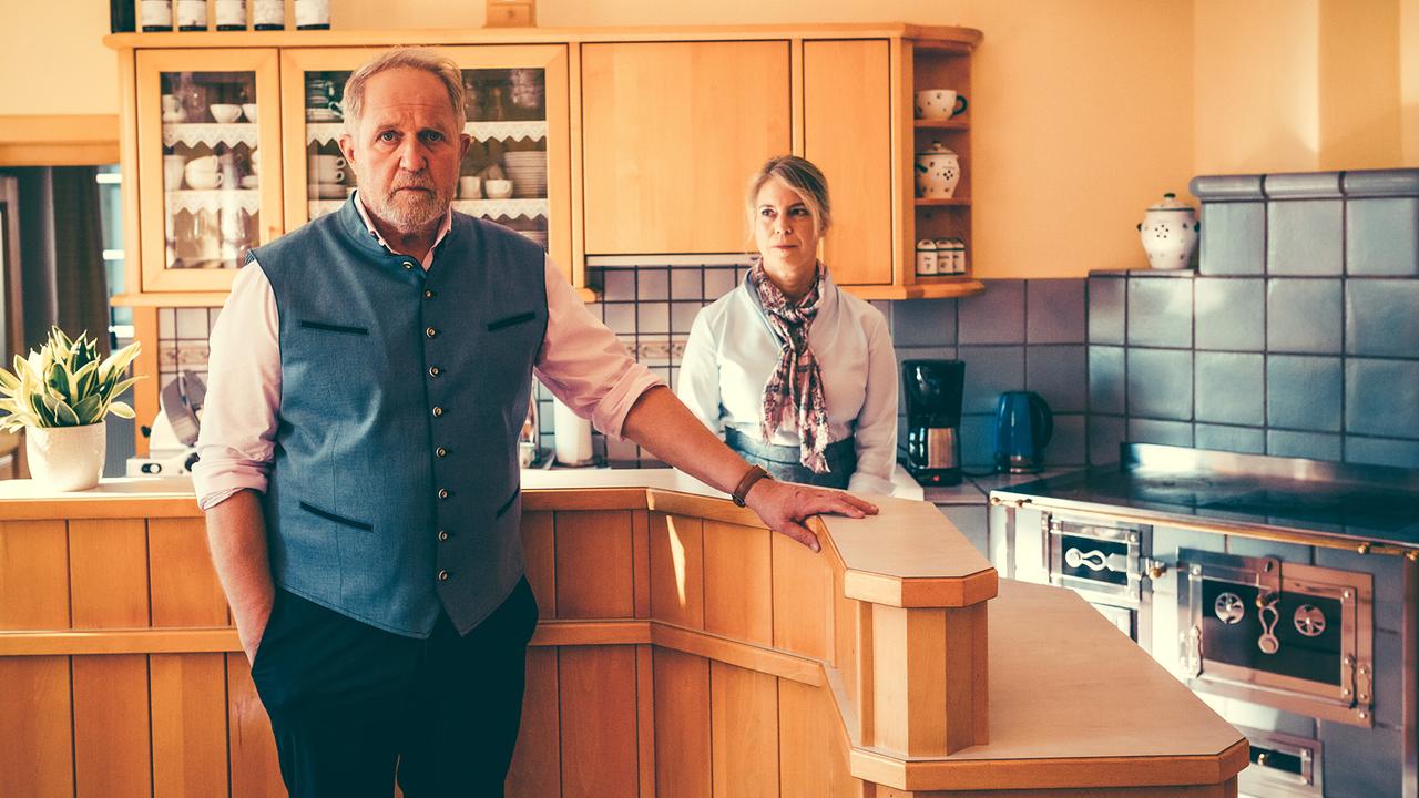 Harald Krassnitzer und Elisabeth Krön stehen ernst schauend an einer hölzernen Theke in einer Küche mit traditionellen Elementen.