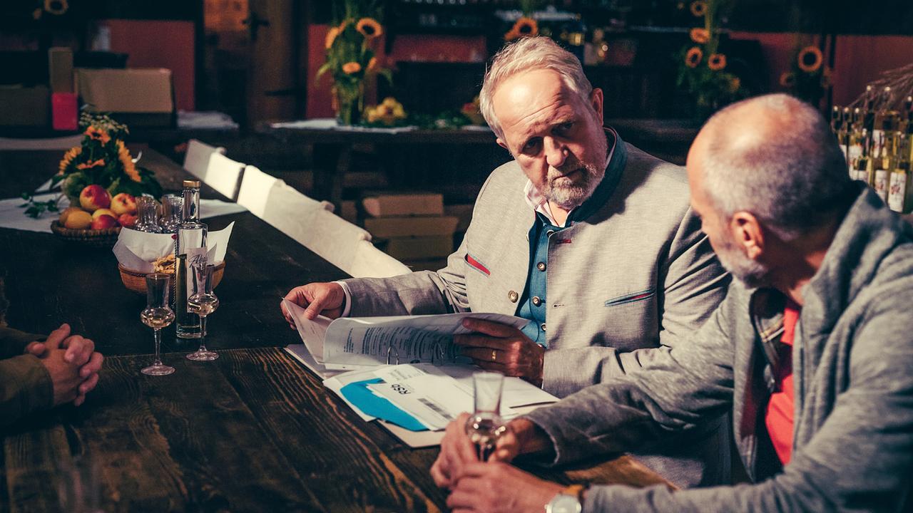 Harald Krassnitzer blättert in Unterlagen und blickt Simon Hatzl an, während  sie an einem rustikalen Holztisch mit Schnapsgläsern sitzen.