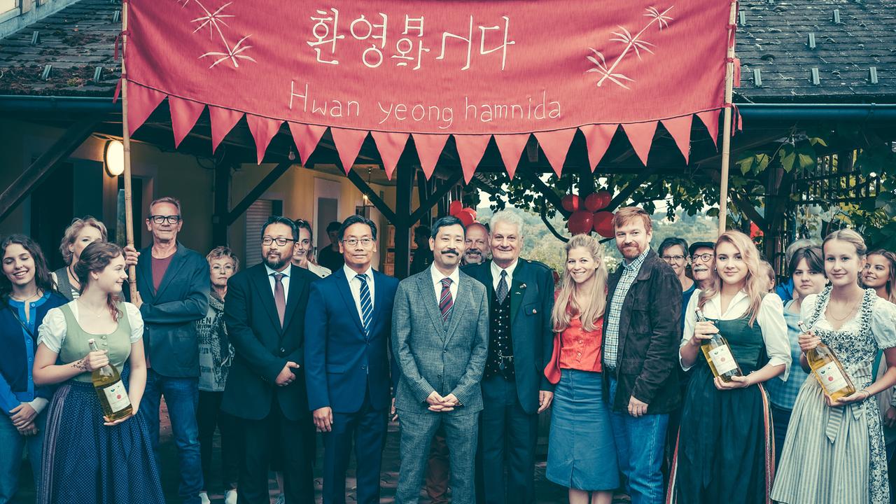 Gruppe vor einem Gebäude unter rotem Banner mit koreanischer Schrift, einige in Tracht halten Weinflaschen in den Händen, andere in Anzügen.