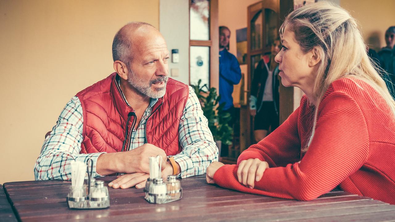 Simon Hatzl und Hilde Dalik sitzen an einem Holztisch im Freien und unterhalten sich, im Hintergrund stehen weitere Personen im Türbereich.