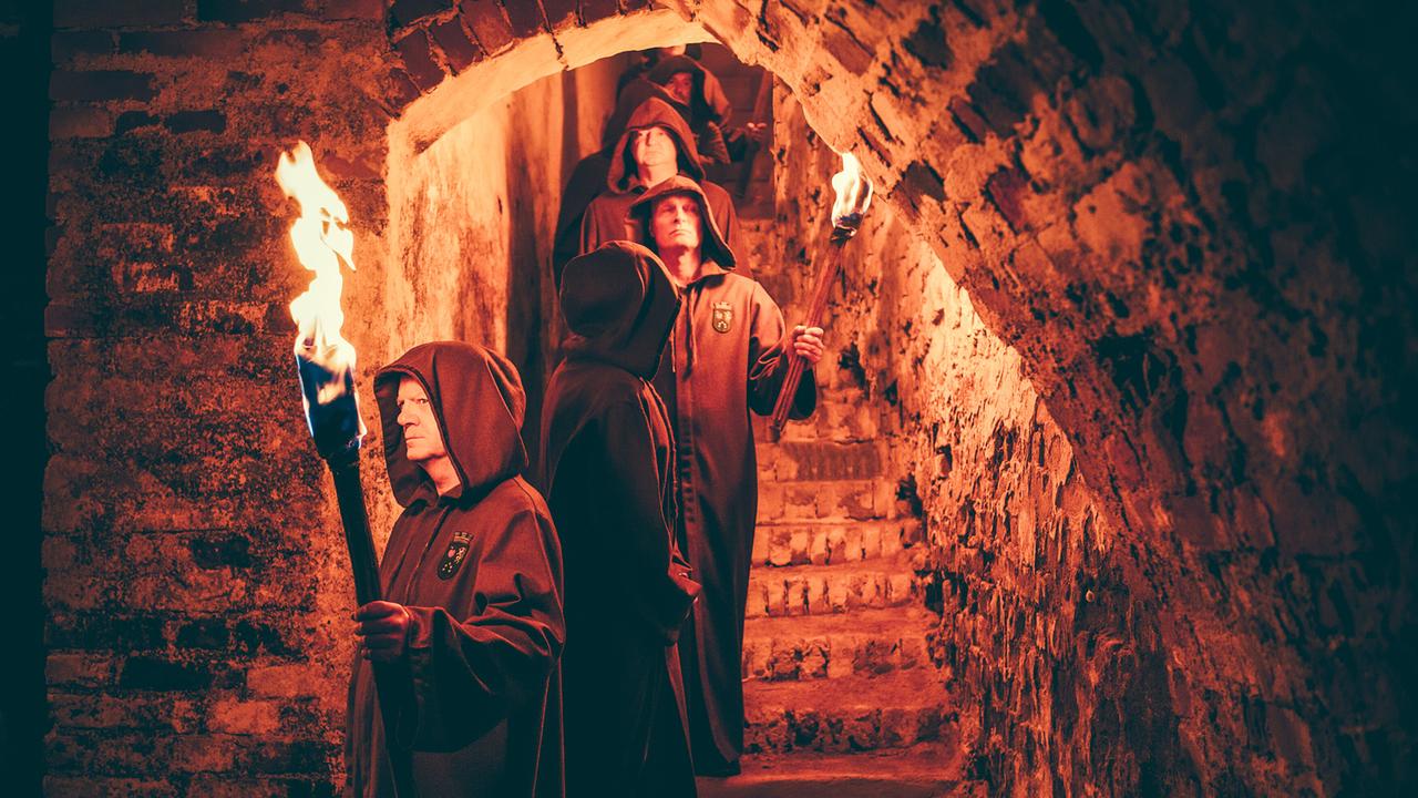 Männer in braunen Kutten mit Fackeln gehen eine steinerne Treppe in einem Gewölbe hinab.
