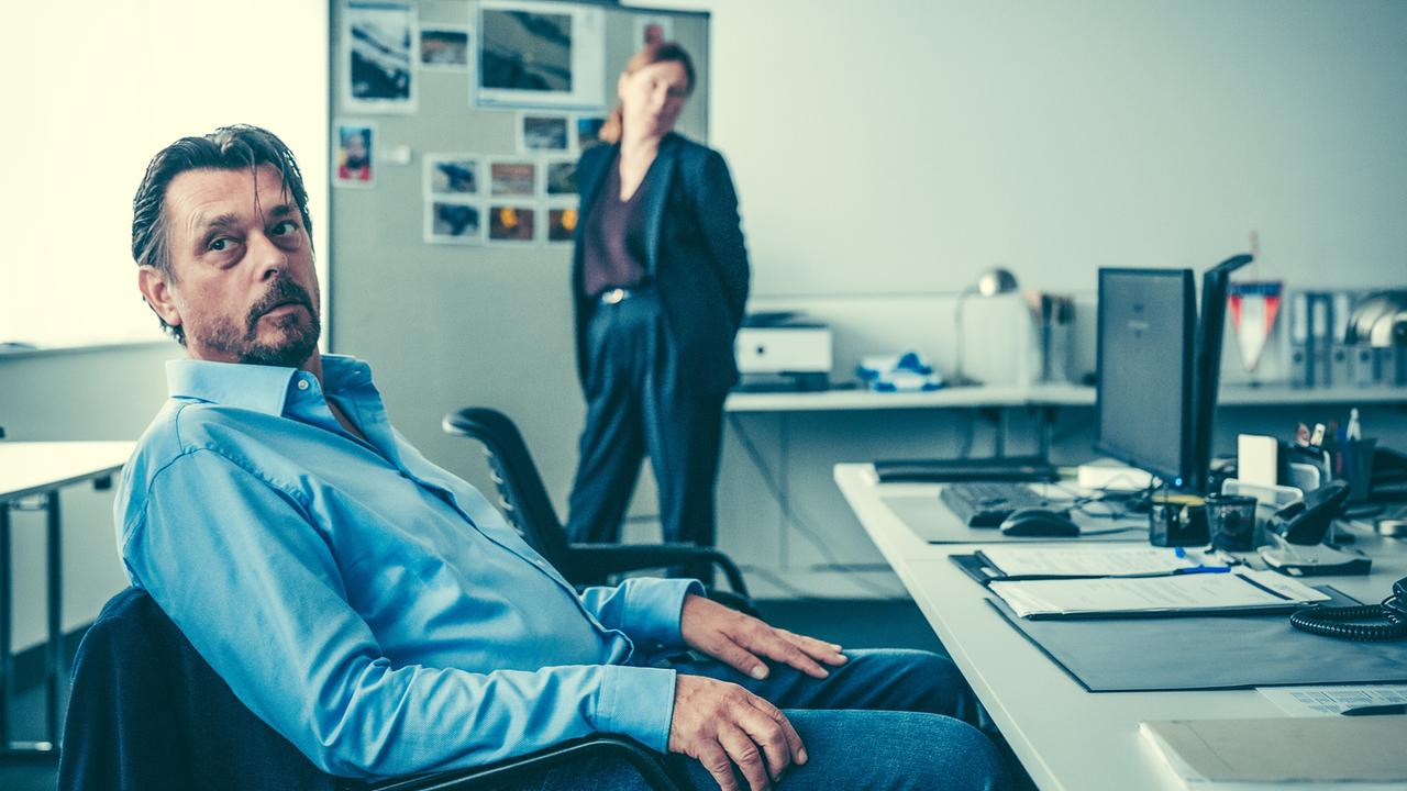 Hary Prinz sitzt in einem Büro an einem Schreibtisch,  im Hintergrund steht Bettina Mittendorfer vor einer Pinnwand mit Fotos.
