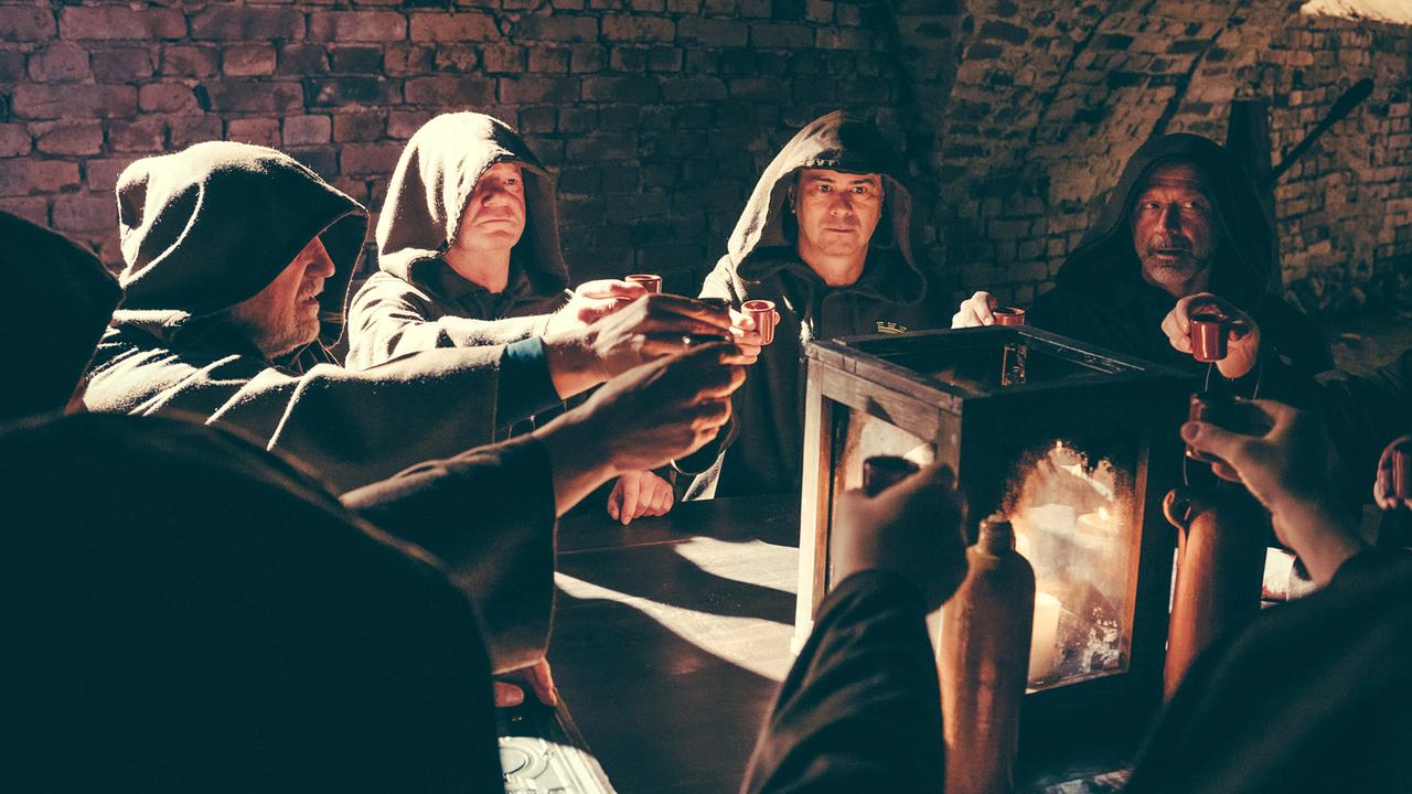 Mehrere Personen unter anderem Harald Krassnitzer in Kutten stoßen mit kleinen Bechern an einem Tisch in einem Gewölbe an.
