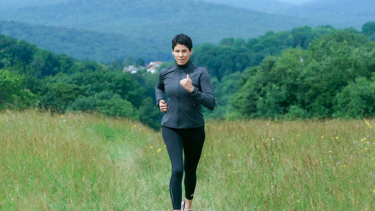 © ORF/MR Film/Petro Domenigg Jasmin Gerat joggt auf einem Wiesenweg mit bewaldeten Hügeln im Hintergrund.