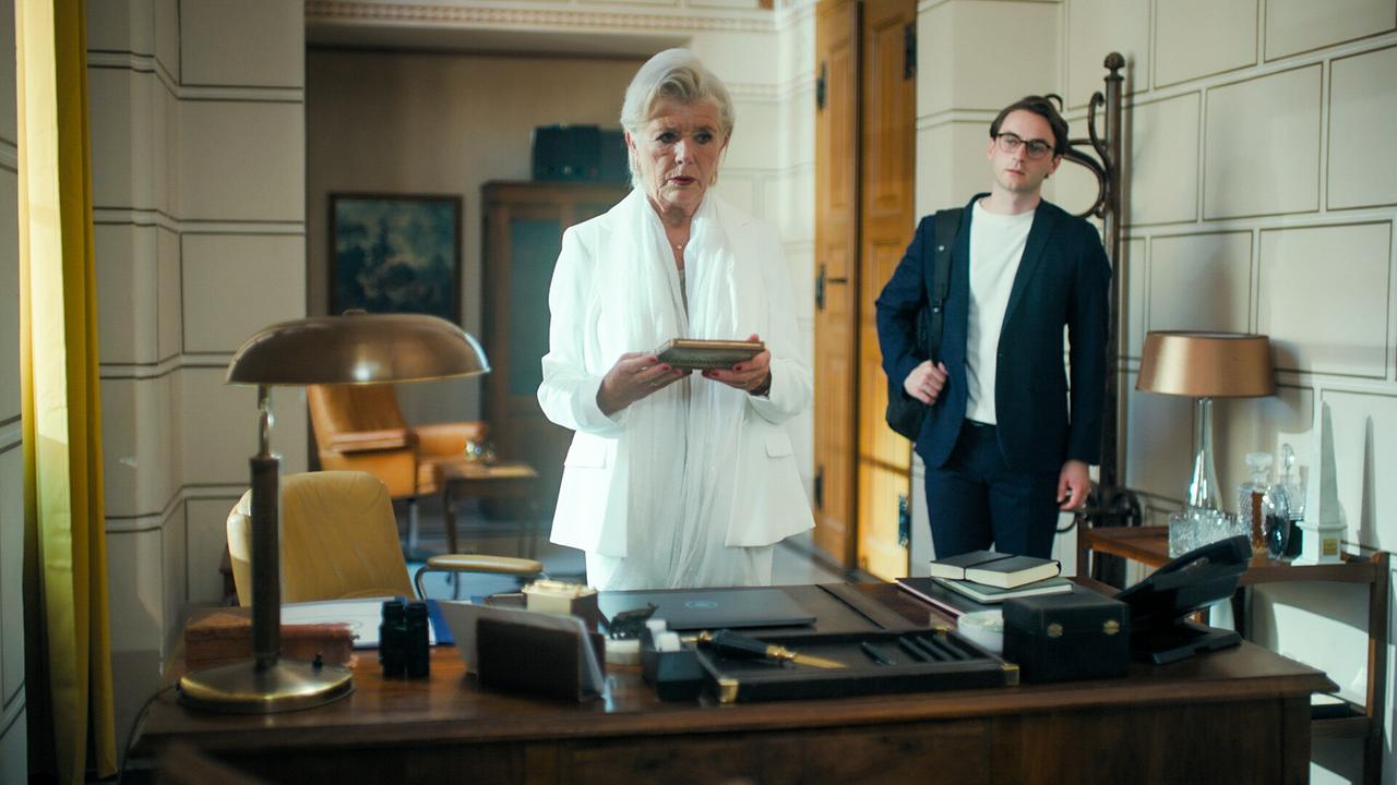 © ORF/MR Film Jutta Speidel steht an ihrem Schreibtisch im Büro und hält ein Buch in der Hand, Tobias Resch steht hinter ihr.