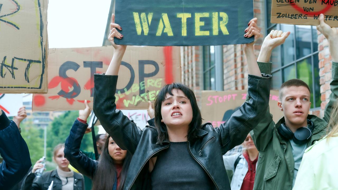 © ORF/MR Film/Petro Domenigg Paulina Hobratschk in schwarzer Lederjacke hält bei einer Demonstration ein Schild mit der Aufschrift „WATER“ hoch.