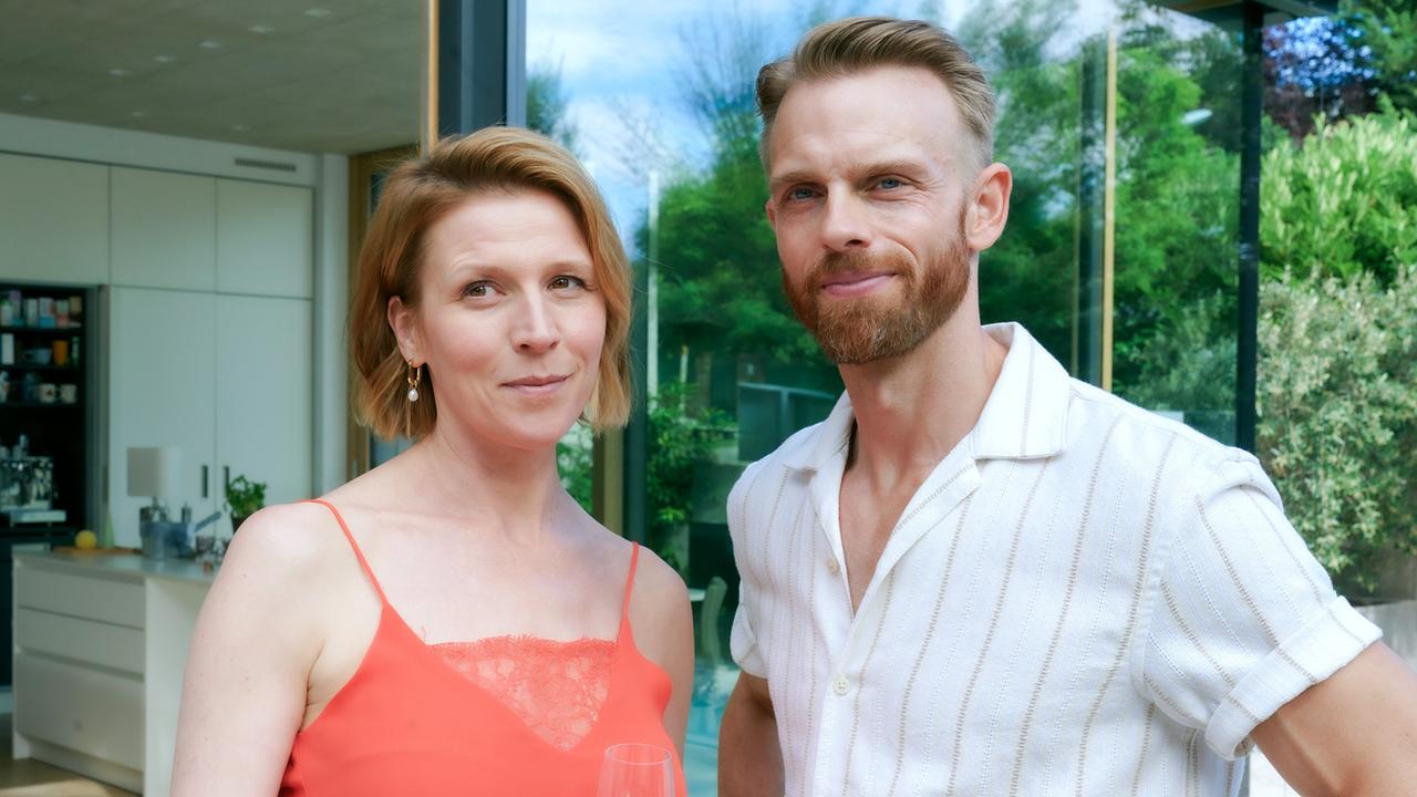 © ORF/MR Film/Petro Domenigg Franziska Weisz im roten Top steht neben Mads Hjulmand im kurzärmeligen Hemd und hält ein Glas Sekt.