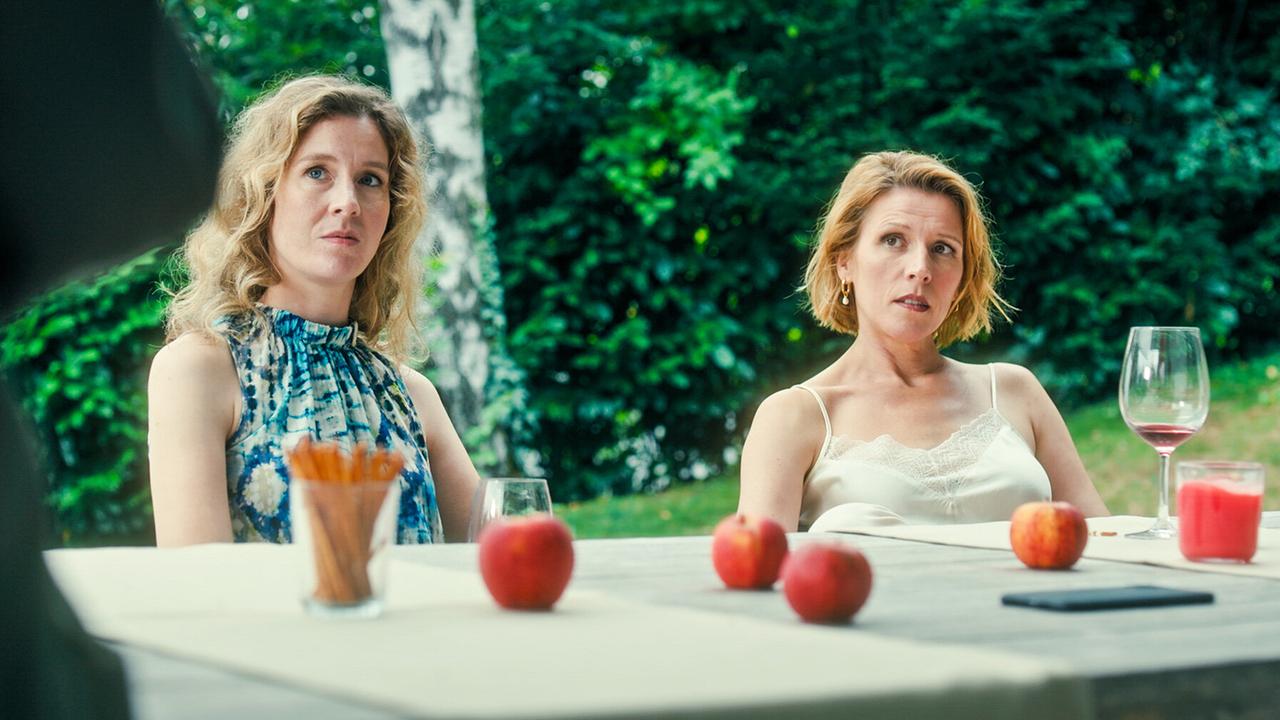 Franziska Hackl und Franziska Weisz sitzen im Freien an einem Gartentisch mit Getränken, roten Äpfeln und Snacks, im Hintergrund Bäume und Grün.