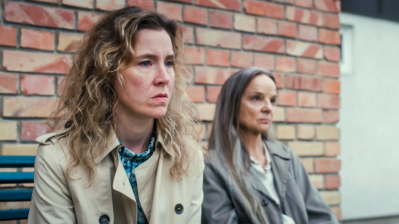 Franziska Hackl und Sissy Höfferer sitzen auf einer Bank vor einer Ziegelwand und schauen ernst.