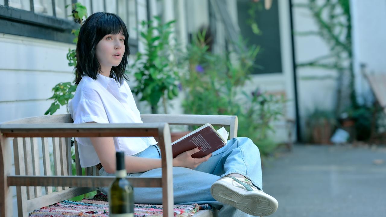 Paulina Hobratschk sitzt draußen auf einer Holzbank, liest ein Buch und hat eine Weinflasche neben sich stehen.