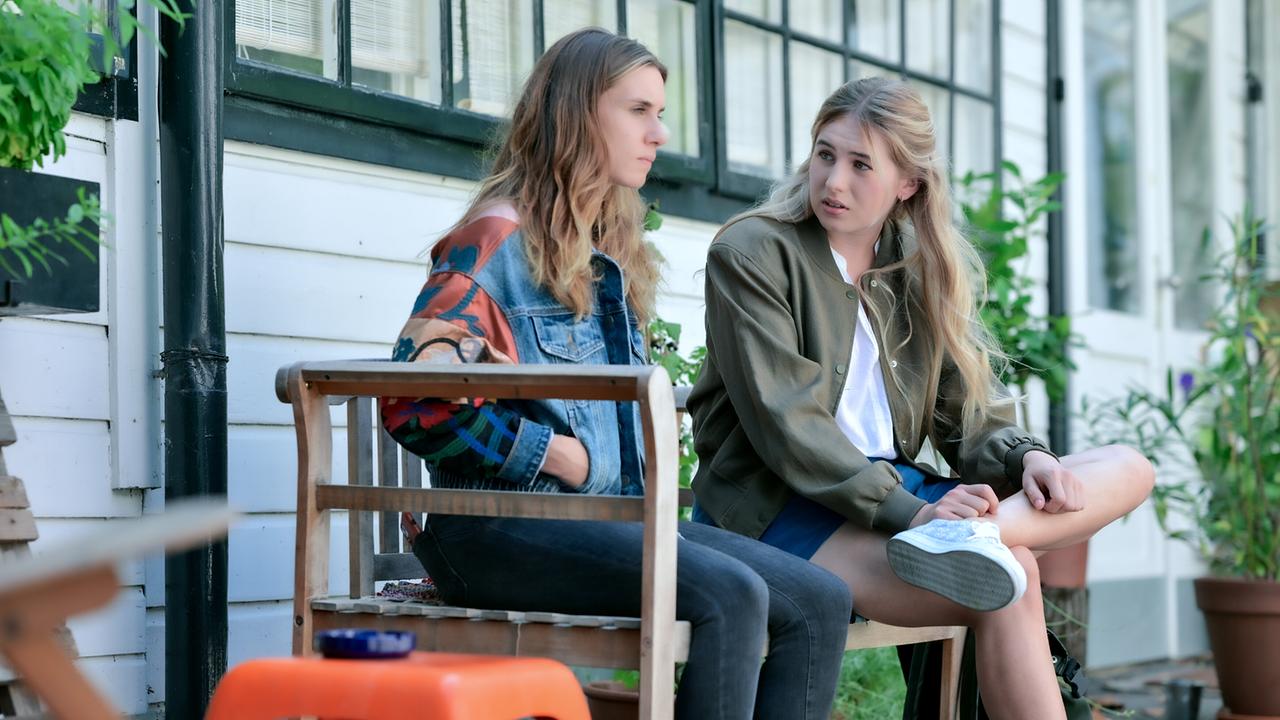 Maya Unger und Niobe Eckert sitzen im Gespräch auf einer Holzbank vor einem Gebäude mit großen Fenstern.