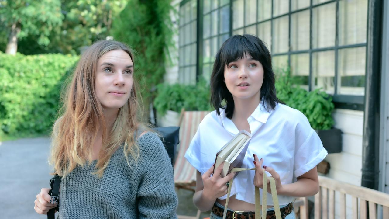 Maya Unger und Paulina Hobratschk, die ein Buch in der Hand hält, stehen nebeneinander vor einer Fassade aus horizontalen, weiß gestrichenen Holzpaneelen mit Sprossenfenstern.