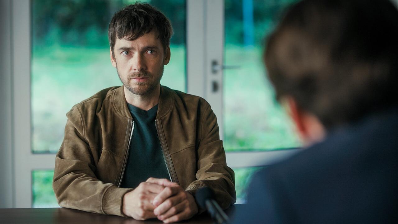 Stefan Pohl sitzt an einem Tisch und spricht mit einem Mann, im Hintergrund große Fenster mit Blick ins Grüne.
