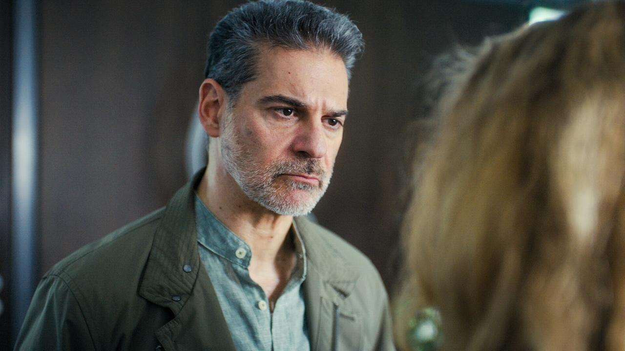 Rick Kavanian blickt in ienem Raum ernst auf eine weitere Person, im Hintergrund eine dunkle Wand.